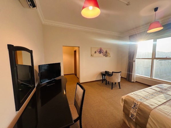 A hotel room with a bed, a desk with a television, a chair, a small table, and a painting on the wall. - Al Jawhara Suite Serviced Apartments 2 (Taif)