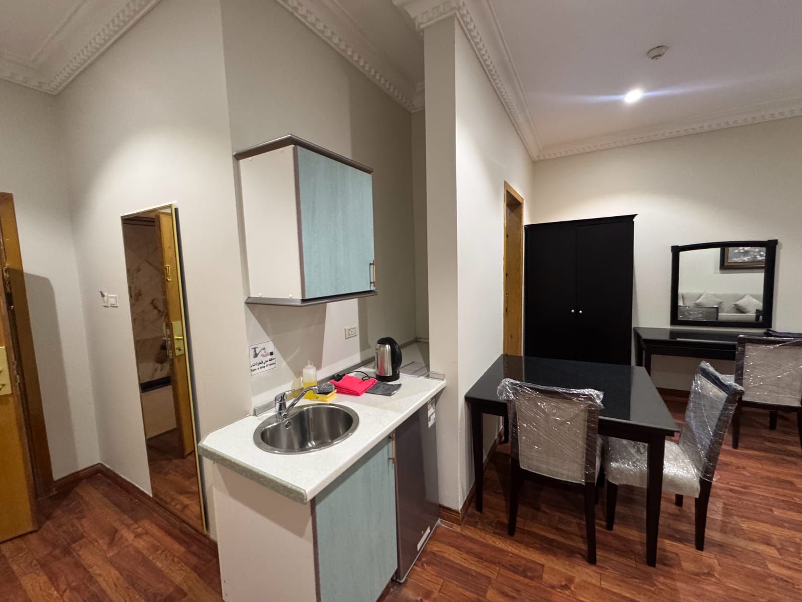 Cozinha compacta com pia, cafeteira e armário. Há uma mesa de jantar com cadeiras, algumas cobertas com plástico.