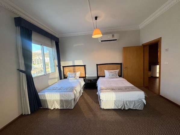 A hotel room with two single beds, a window with curtains, an air conditioning unit, and a door leading to another room.