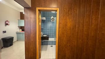 Een betegelde badkamer met houten lambrisering, een douchegedeelte met blauwe tegels, een zichtbare wastafel en een deur naar een gang.