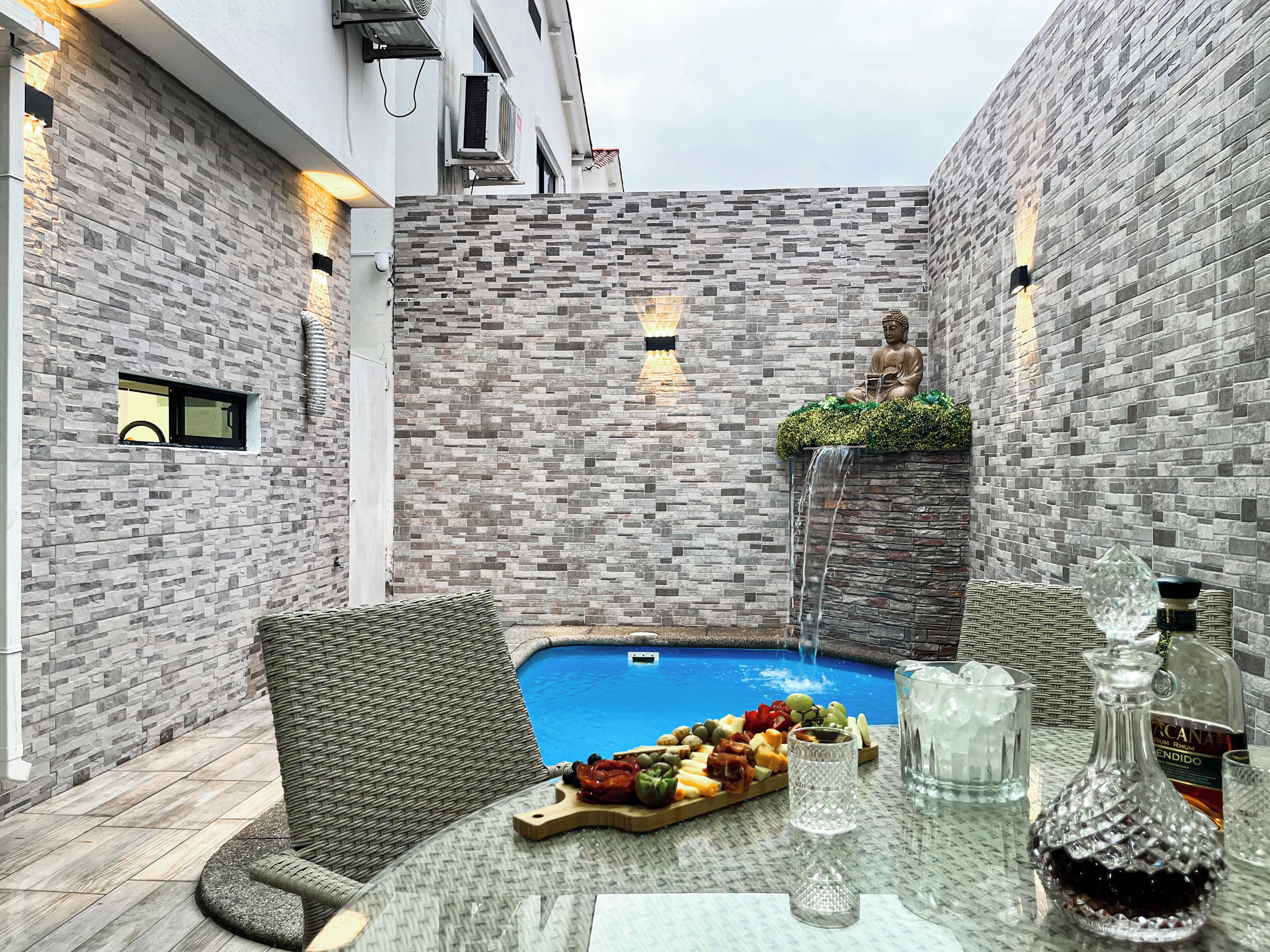 A poolside area with a statue, a wicker chair, a table with a fruit platter, and a bottle of liquor.