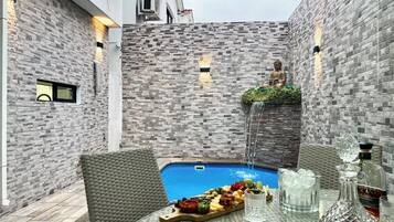 A poolside area with a statue, a wicker chair, a table with a fruit platter, and a bottle of liquor.