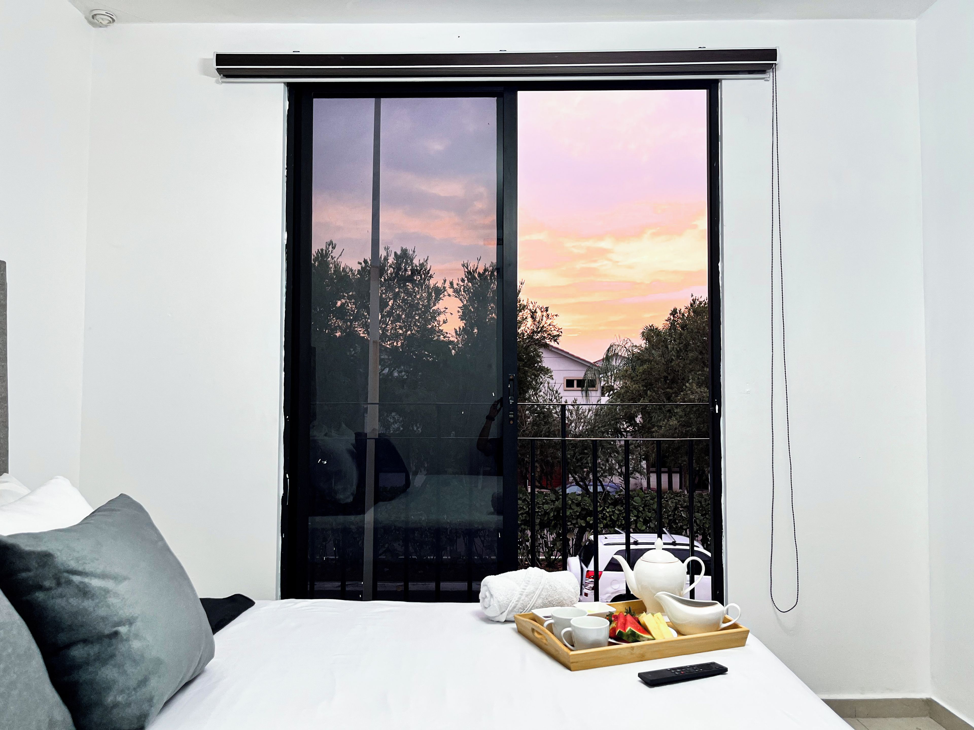 A bedroom with a bed, a bedside table with a tray of breakfast items, a remote control, and a view of trees and a house through a sliding door.