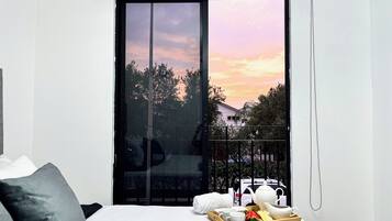A bedroom with a bed, a bedside table with a tray of breakfast items, a remote control, and a view of trees and a house through a sliding door.