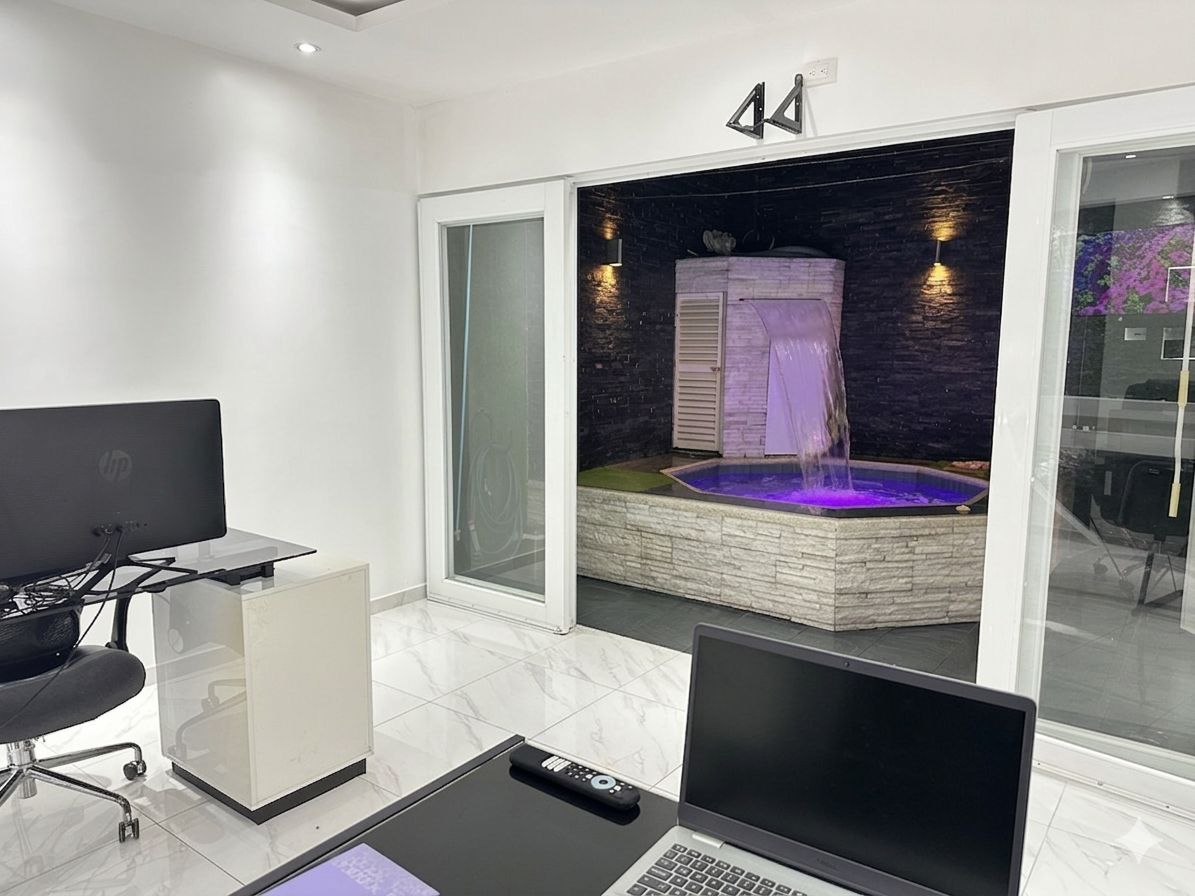 A modern room with a large window, a laptop, a remote control, and a spa area with a hot tub and waterfall.