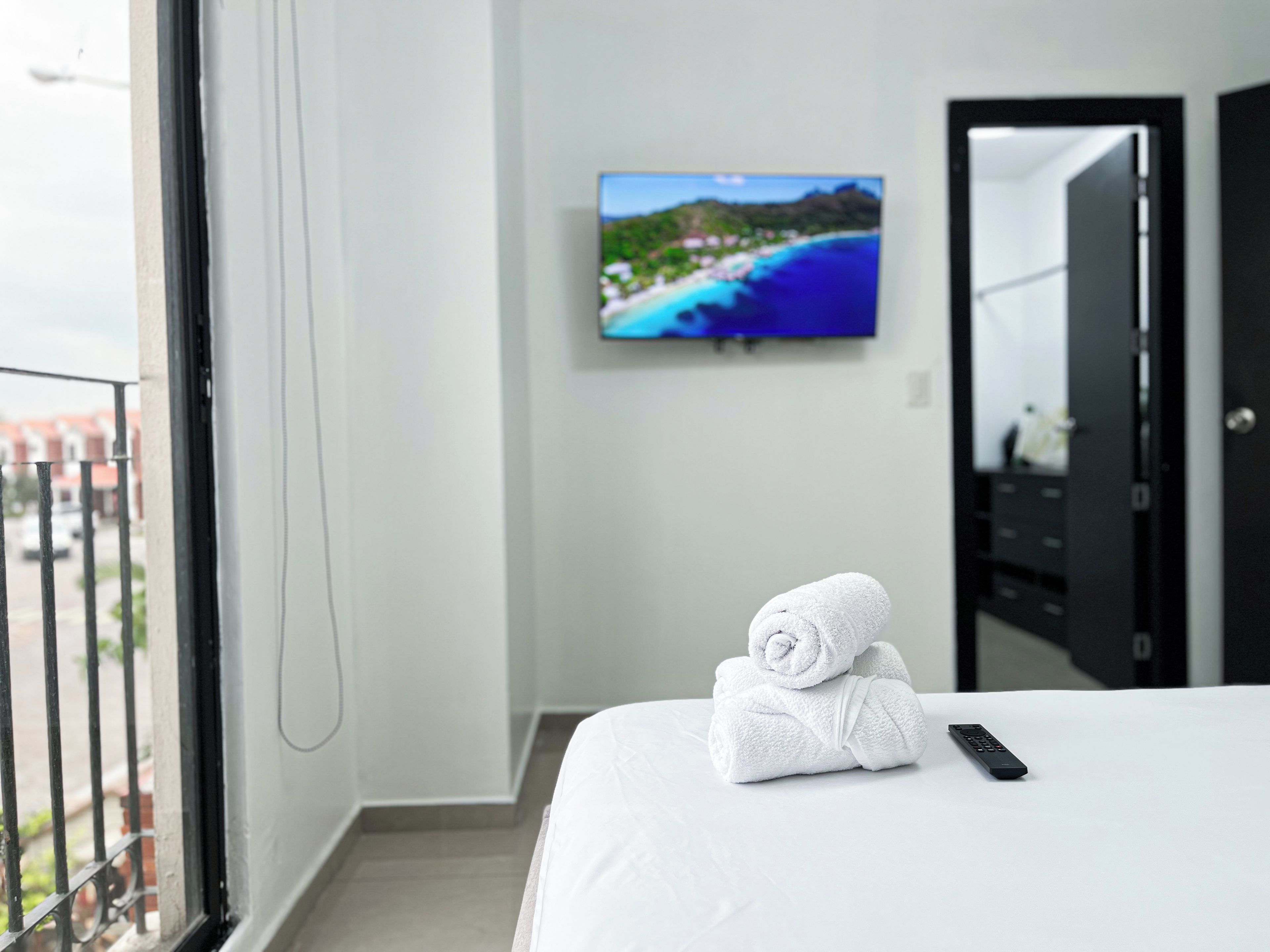 Camera d'albergo con un letto, un asciugamano piegato, un telecomando e una televisione a parete che mostra una veduta costiera.