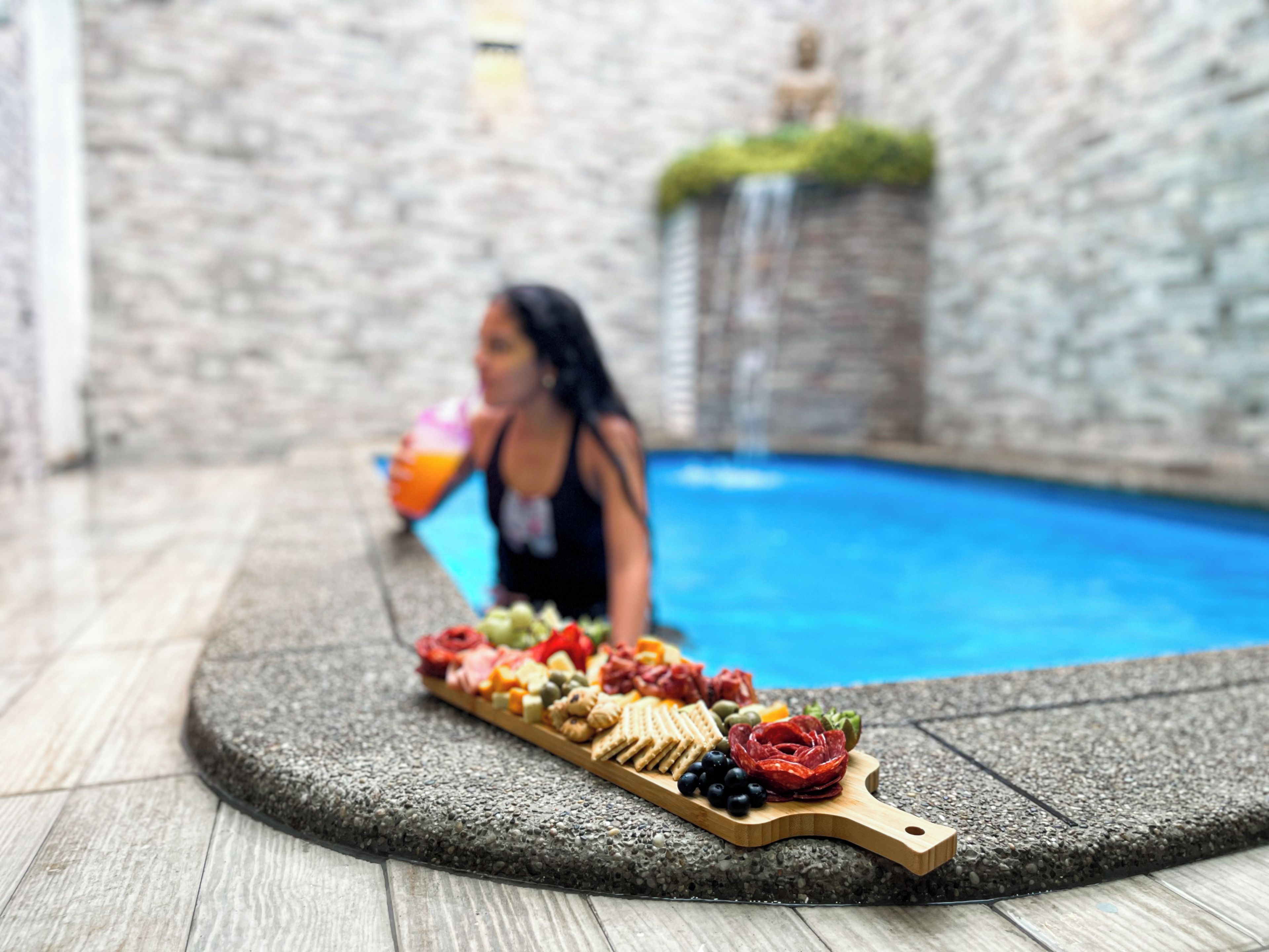 A poolside area with a wooden board displaying an assortment of snacks and a person holding a drink.