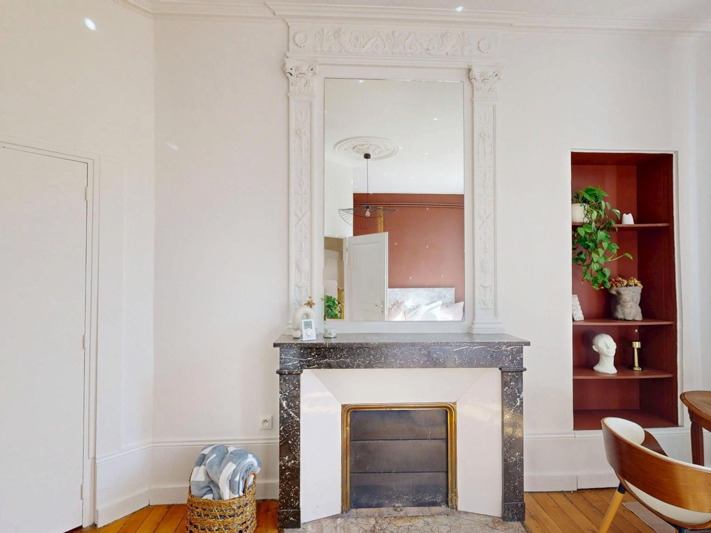A room with a fireplace, a mirror, a wooden floor, a chair, and a shelf with decorative items.