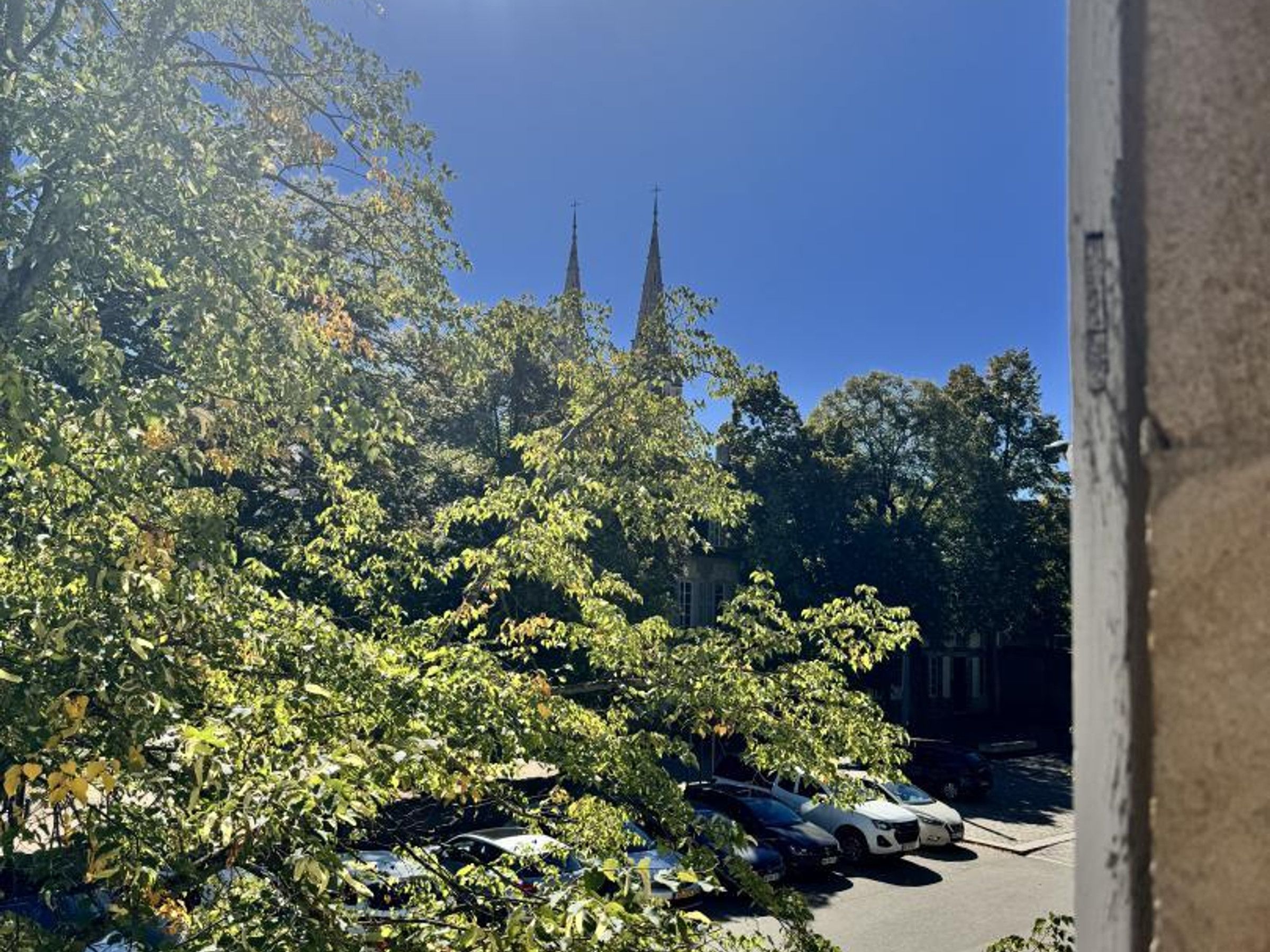Un parking avec des voitures, des arbres et un bâtiment surmonté d’une haute flèche, le tout sur fond de paysage.