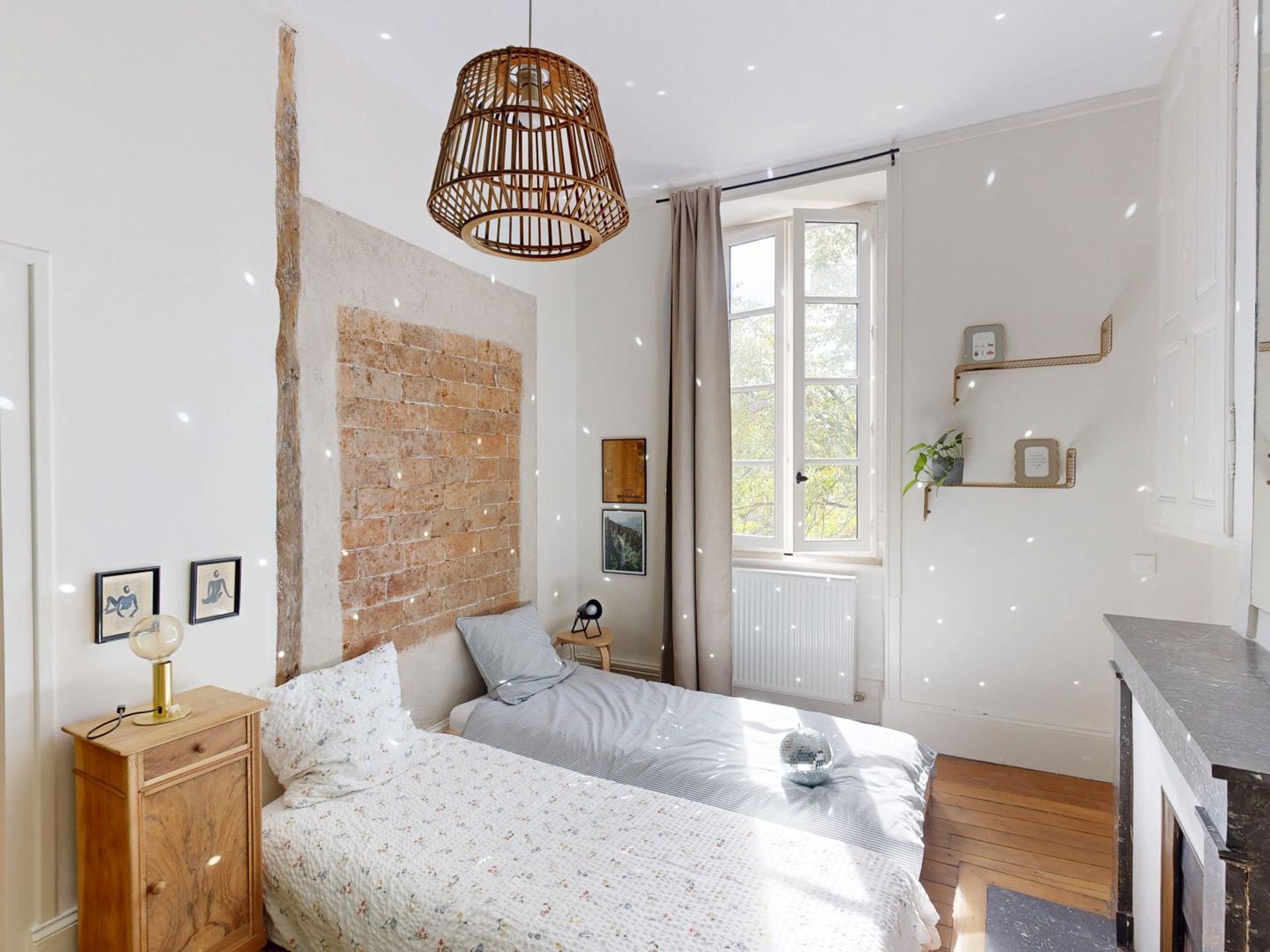 A bedroom with two beds, a brick wall, a hanging light fixture, and a window with curtains.