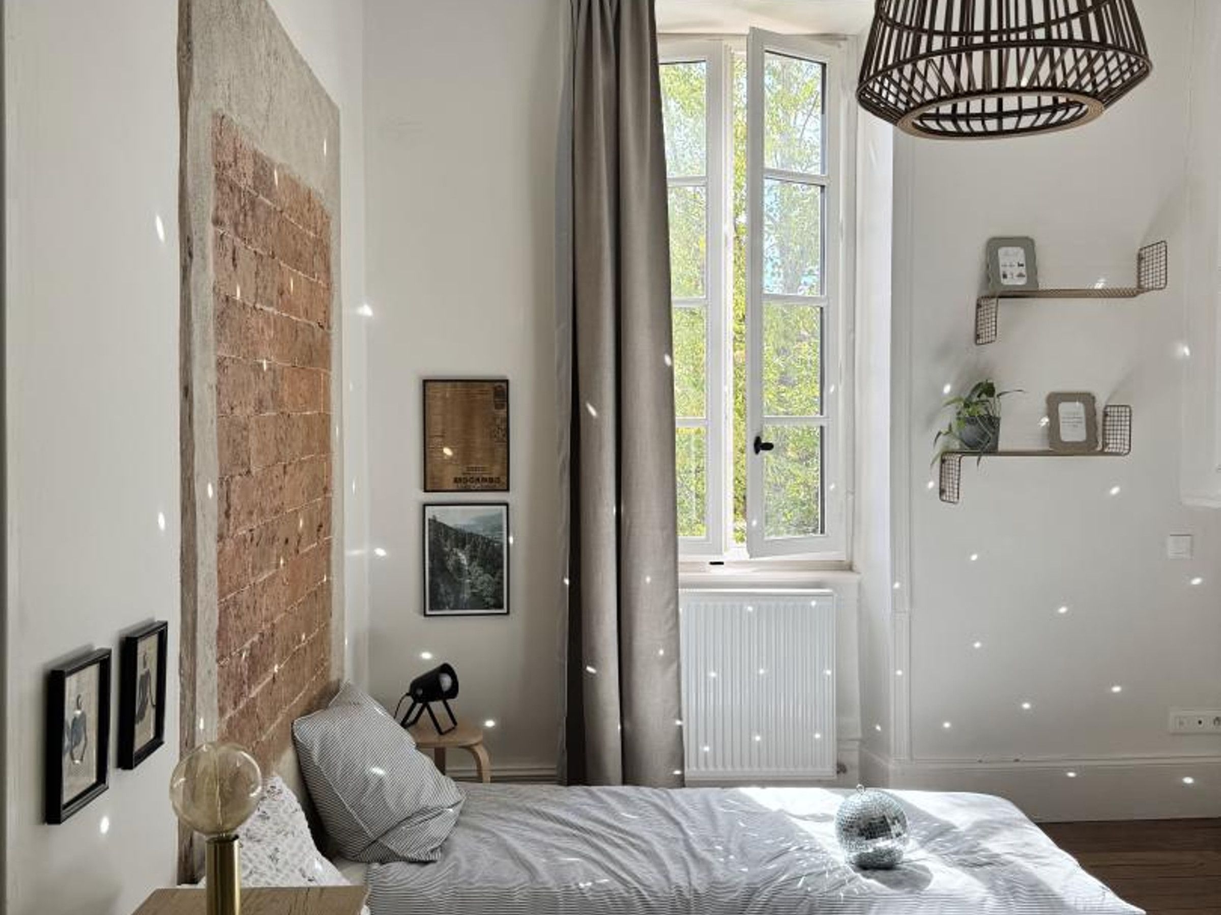 Een slaapkamer met een bakstenen muur, een bed met een gestreept dekbedovertrek, een hanglamp en een raam met uitzicht op groen.