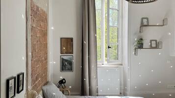 Een slaapkamer met een bakstenen muur, een bed met een gestreept dekbedovertrek, een hanglamp en een raam met uitzicht op groen.