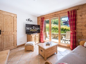A room with a television, wooden furniture, a sofa, and a view of greenery outside. - Spacious apartment with terrace and pool (Les Menuires)