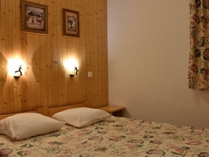 A room with a wooden paneled wall, two wall-mounted light fixtures, a bed with floral bedding, and a small wooden bench. - Cozy apartment mountain view - 5 guests (Méribel)