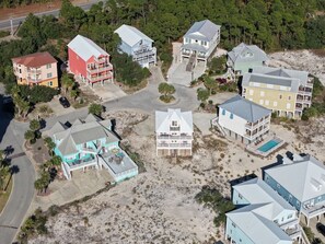 Vue aérienne d’un quartier résidentiel situé sur la côte, avec de nombreuses maisons à deux étages, dont certaines sont dotées de balcons, et une plage de sable en arrière-plan.