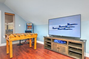 A room with a foosball table, a flat-screen TV displaying an airplane, and a shelf unit. - Flip Oasis Lockout · Coastal Comfort: Hot Tub, Foosball & Arcade (Gulf Shores)
