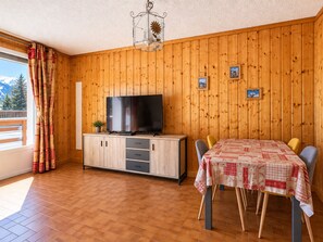 A room with a wooden paneled wall, a television on a cabinet, a dining table with chairs, and a balcony with curtains. - QUIR301 - Profitez d’un séjour au calme, à deux pas des pistes (Mont-de-Lans)