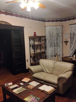 Een woonkamer met een leren bank, een houten salontafel met boeken, een boekenkast en een plafondventilator.