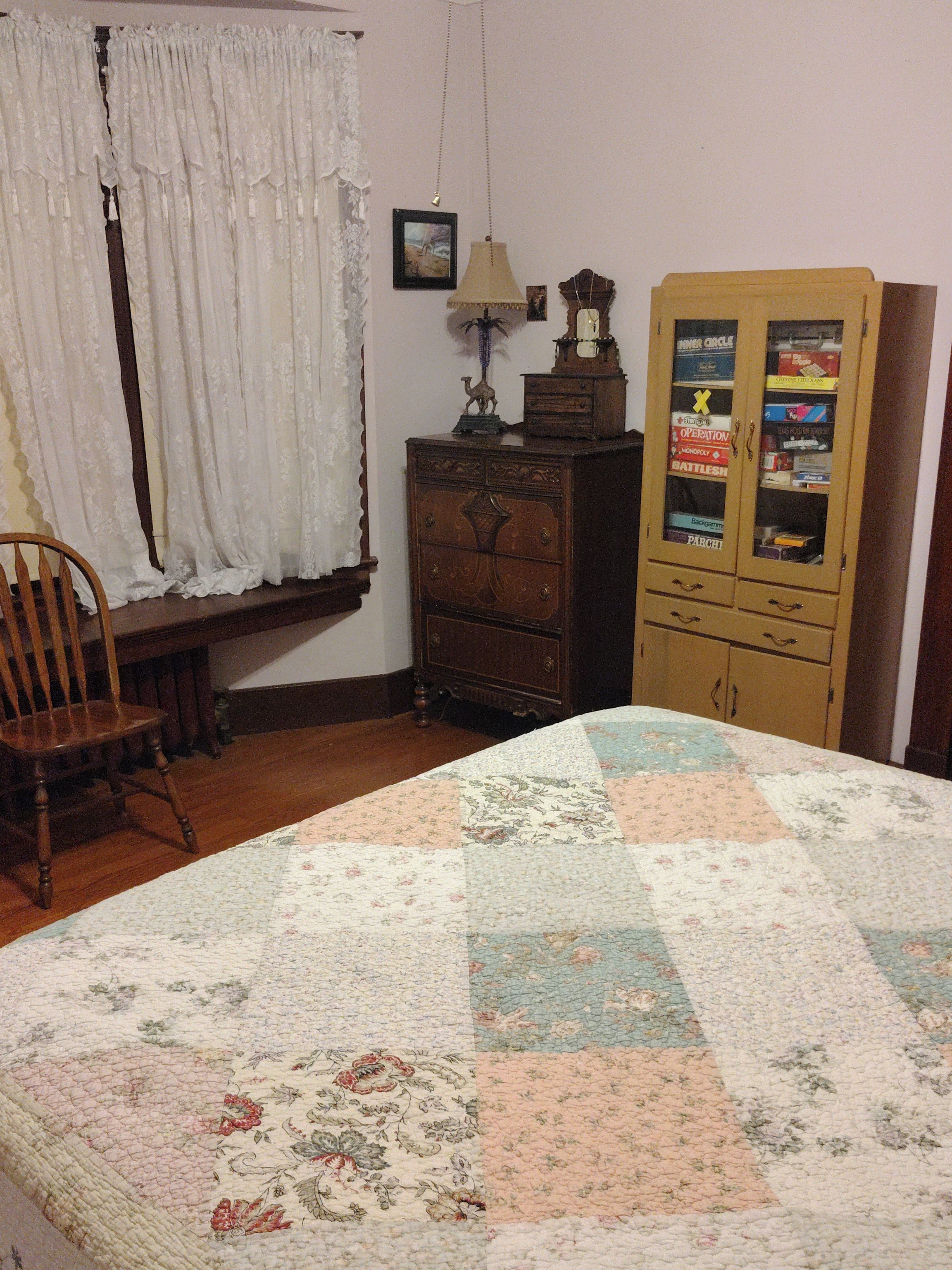 A room with a bed, a wooden cabinet with glass doors, a chair, and a window with curtains.