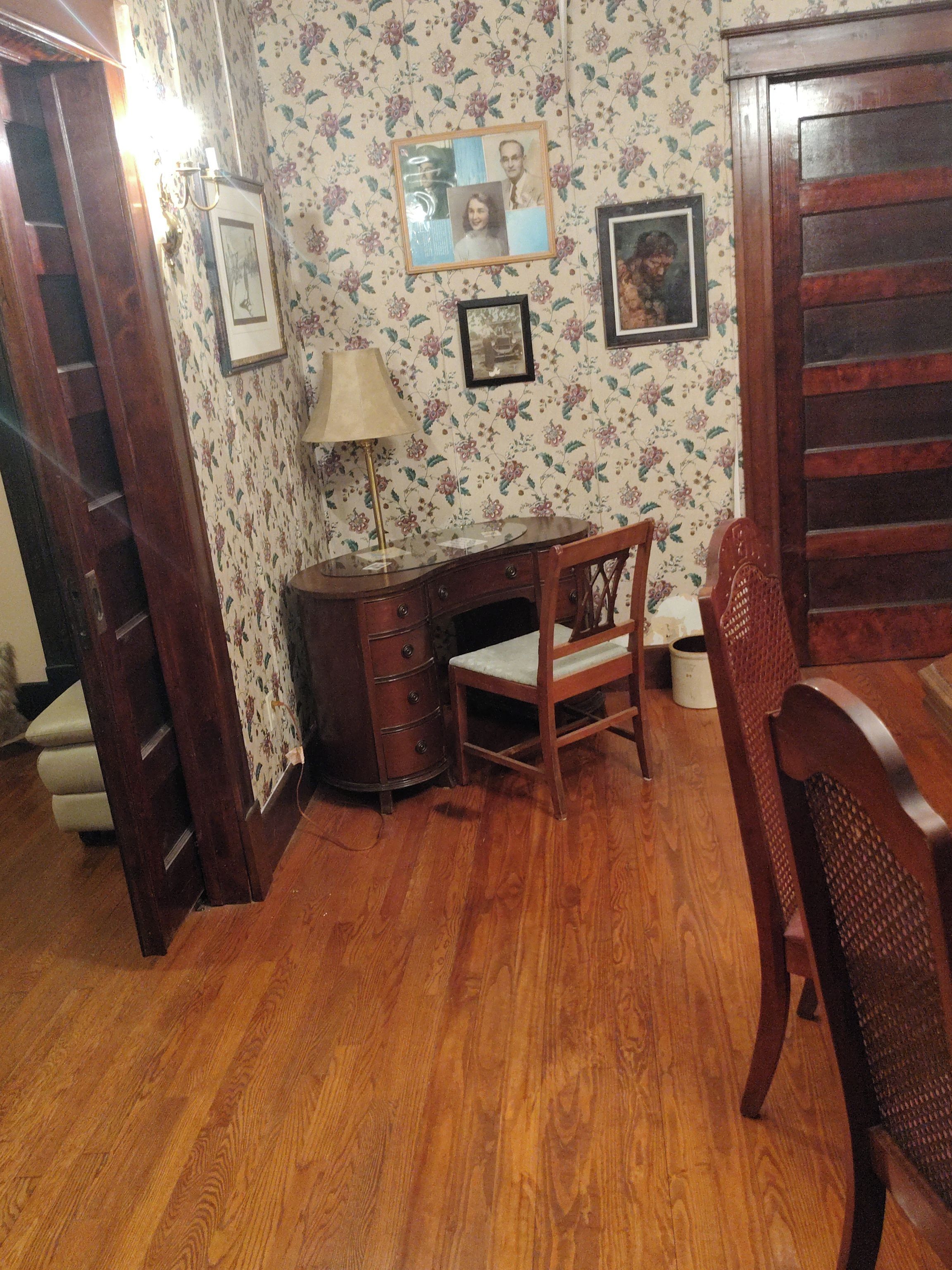 A room with wooden flooring, a desk with drawers, a chair, and a wall with floral wallpaper.