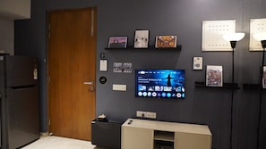 A room with a television displaying a program, a wooden door, and a shelf with framed pictures. - London Studio I Hiranandani Estate I WiFi (Thane)