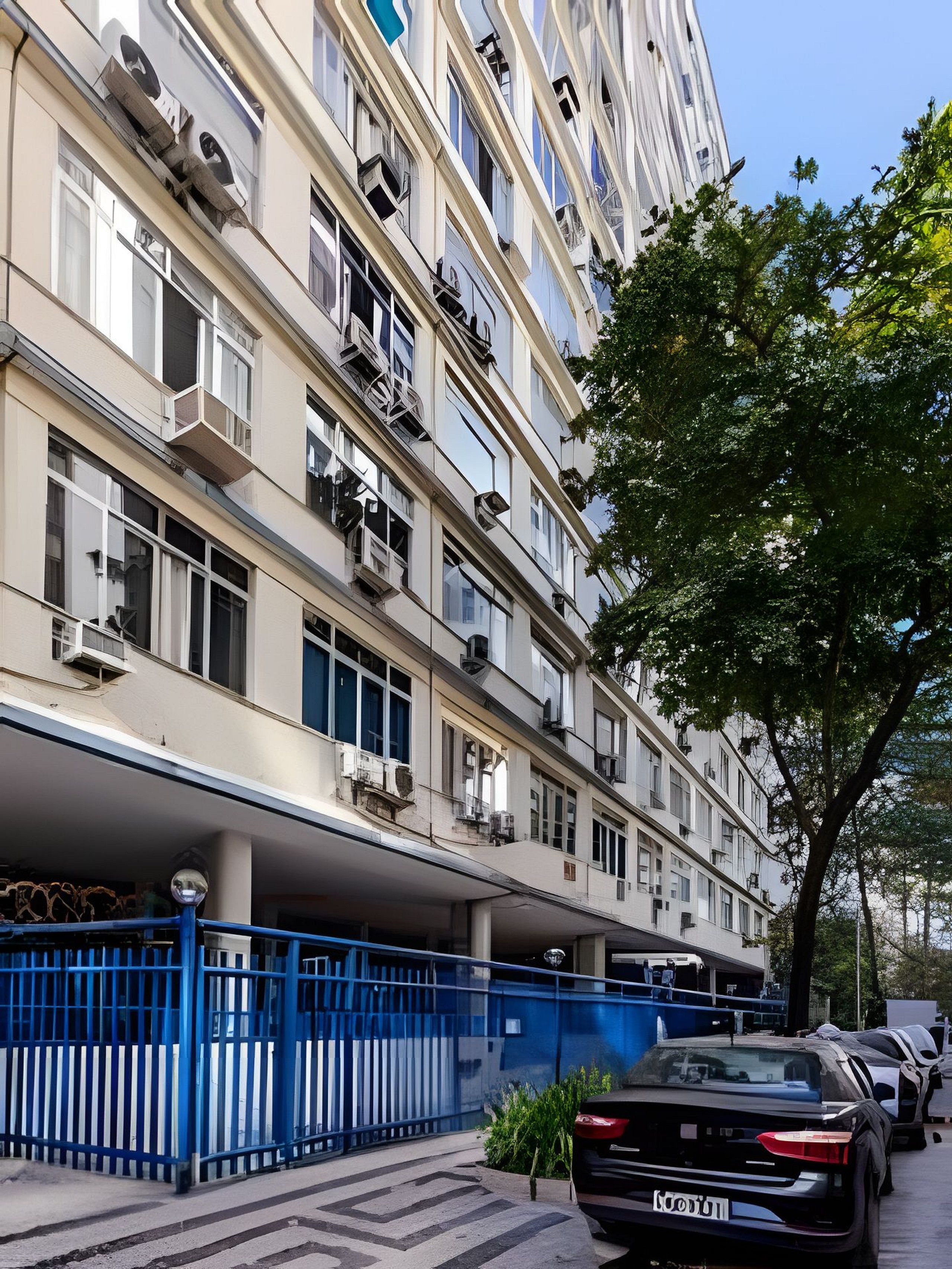 A multi-story residential building with balconies, a blue fence, and parked cars.