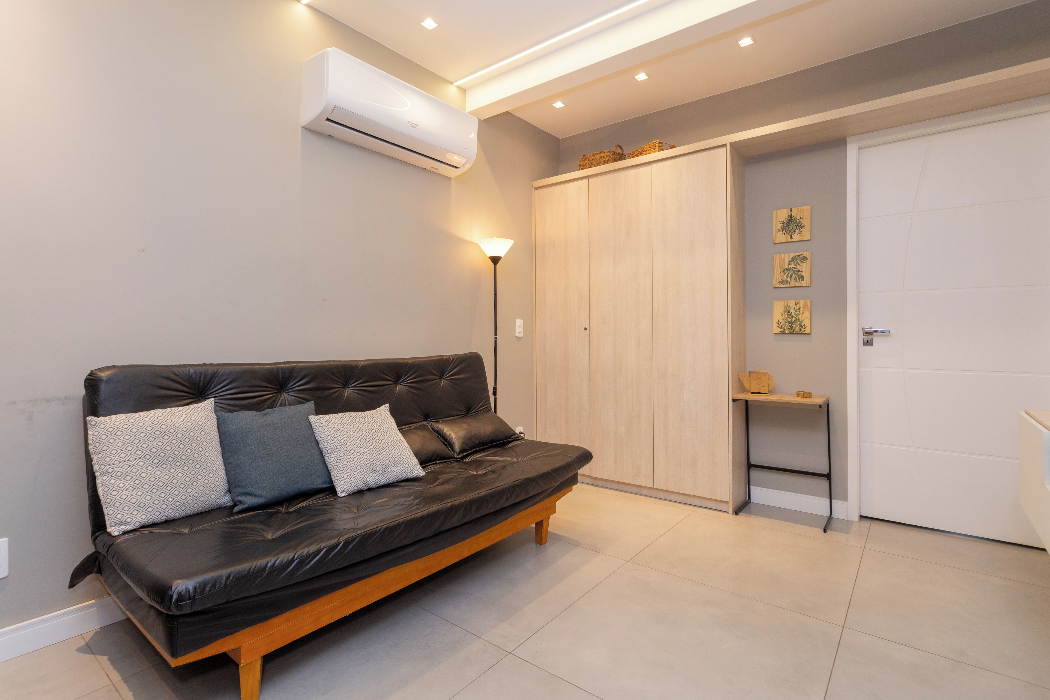 A modern living room with a black leather sofa, a wall-mounted air conditioner, and a wooden wardrobe with a shelf.