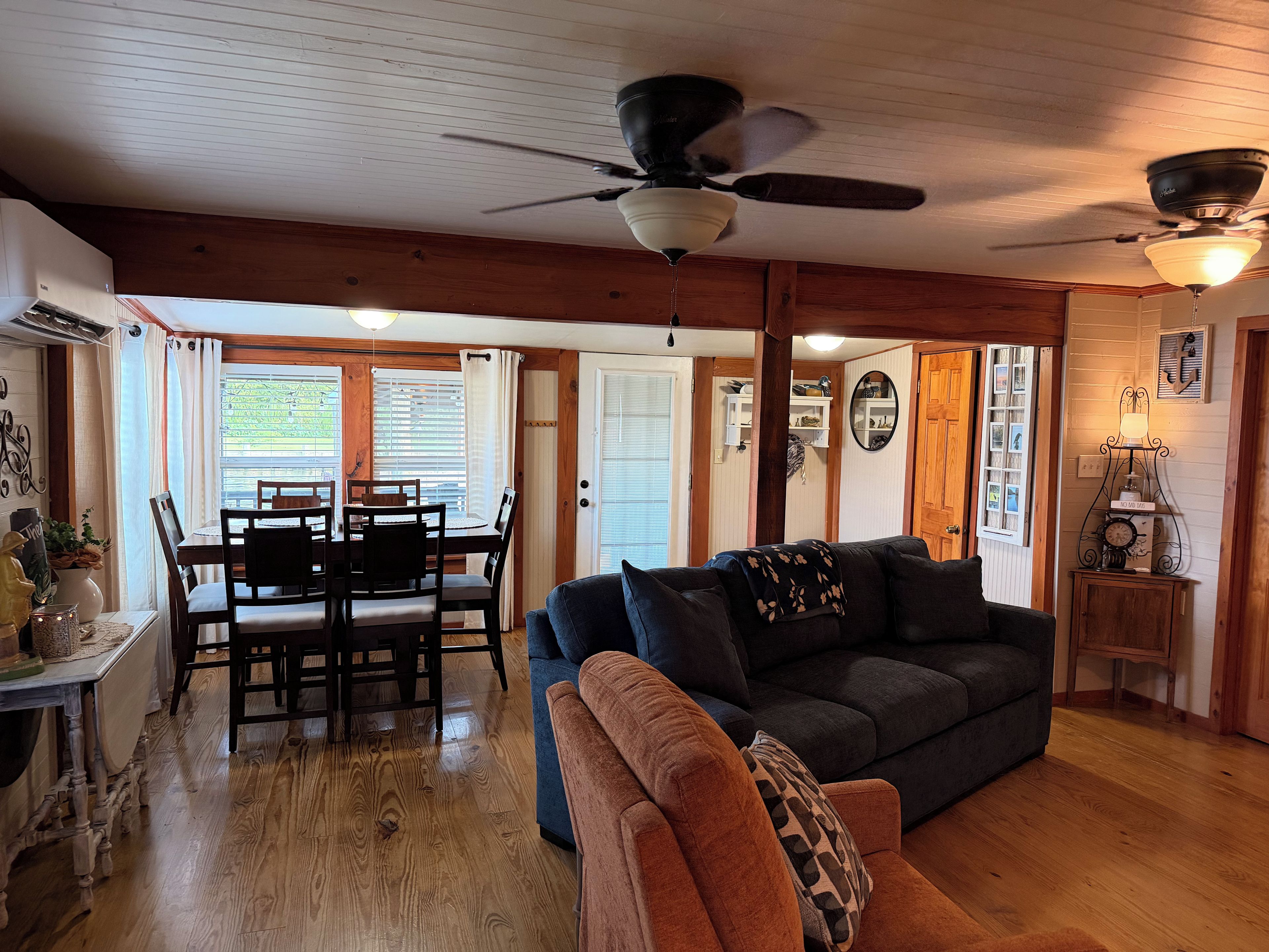 A cozy living room with a wooden floor, a dark-colored sofa, a dining table with chairs, and a ceiling fan.