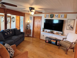 A cozy living room with a dark-colored sofa, a wooden floor, a ceiling fan, a mounted flat-screen TV, and a wooden shelf with decorative items. - The Fisherman’s Hideaway (Vacherie)