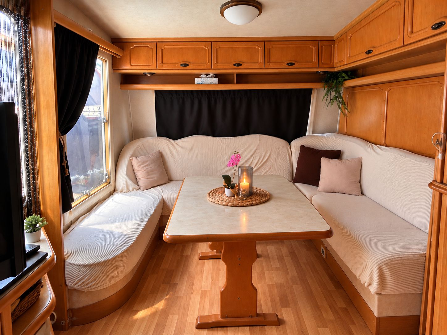 Een gezellig interieur van een camper, met een zachte zithoek, een klein tafeltje met een kaars en een raam met gordijnen.