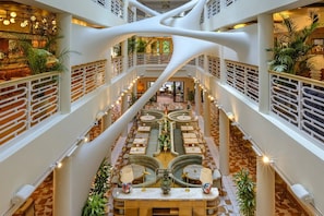 Een meerlaags atrium met een centrale zithoek, omringd door tafels en stoelen, en een modern architectonisch ontwerp met witte pilaren en een patroon vloer.