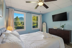 A bedroom with a bed, a dresser, a television, and windows with a view of greenery. - 5BR Smoky Mountain Escape, Hot Tub, Game Room, Sleeps 14,  10 min to Dollywood (Pigeon Forge)