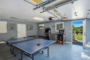 Uma mesa de pingue-pongue em um quarto com uma porta para o exterior, um ar condicionado fixado na parede e uma máquina de jogos eletrônicos.