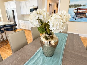 A dining area with a table set for four, a vase with white flowers, and a painting of a boat on the wall. - Bay Breeze Retreat | 6BR Group Getaway | Sleeps 15. (Panama City)