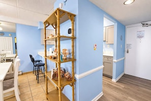 A room with a blue wall, a wooden shelf with decorative items, a dining area with a white table and chairs, and a kitchen area with cabinets. - Atlantis 1403 (Ocean City)