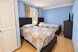 A bedroom with two beds, a window with curtains, a television on a cabinet, and a door with a louvered design. - Atlantis 1403 (Ocean City)