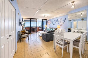 A spacious living area with a dining table, sofa, and chairs. There is a large window providing a view of the outdoors. - Atlantis 1403 (Ocean City)