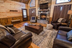A cozy living room with a stone fireplace, leather sofas, and a wooden floor. - Highland Ridge | 4 Bedrooms, 3 King Suites, Private Hot Tub & Fire Pit (Broken Bow)