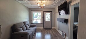 Un couloir avec un canapé gris, une porte ornée d’un panneau de verre décoratif et un plafond agrémenté d’un lustre.