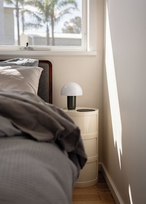 A bedroom with a bed, a bedside lamp, and a window with a view of palm trees.
