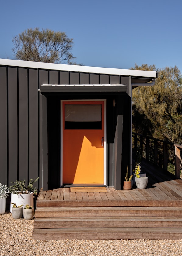 A modern building with a wooden deck, a door with a red frame, and potted plants.