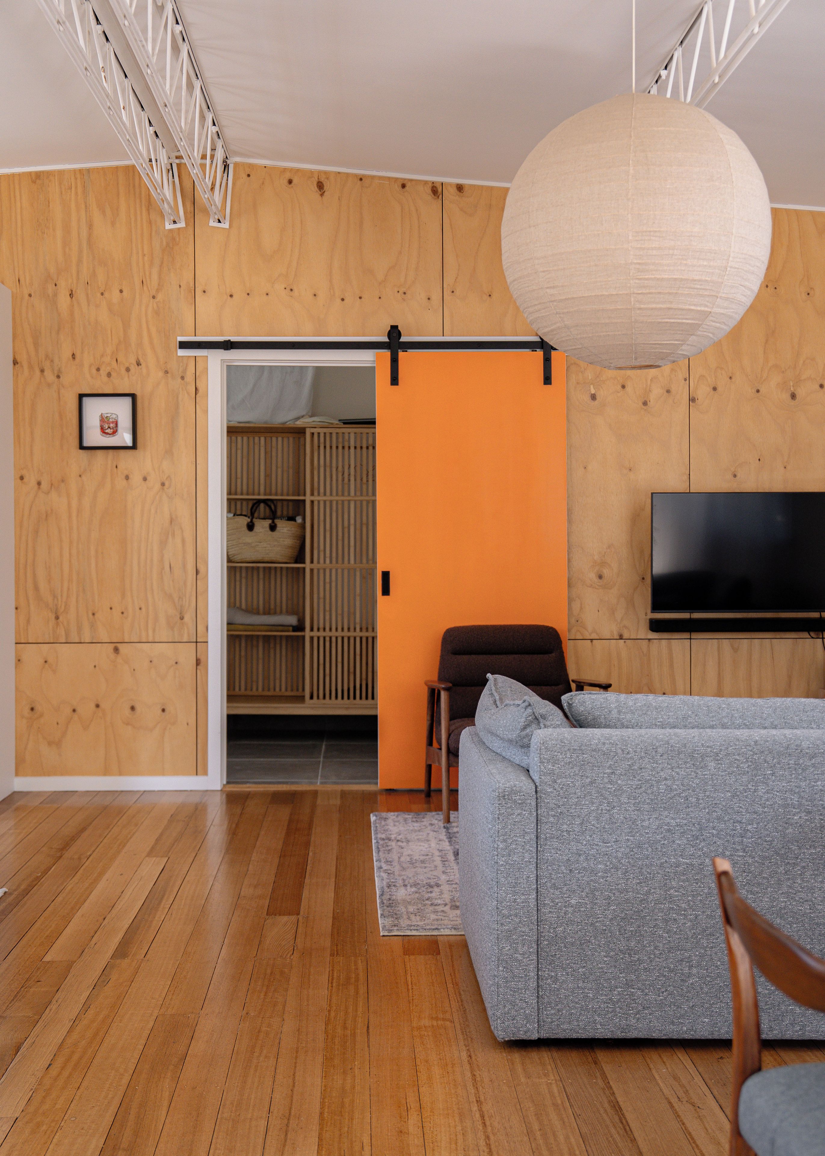 A modern living room with a grey sofa, wooden flooring, a television mounted on the wall, and a sliding door leading to a kitchen area.