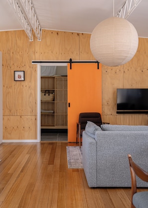 A modern living room with a grey sofa, wooden flooring, a television mounted on the wall, and a sliding door leading to a kitchen area.