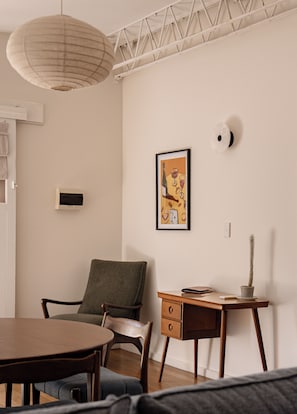 A room with a dining table, a chair, a desk with drawers, a hanging light fixture, and a framed picture on the wall.