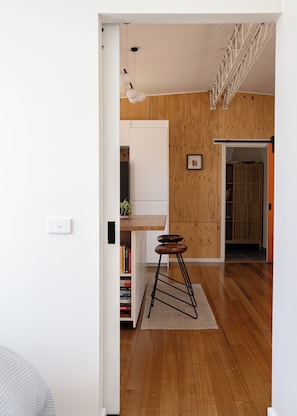A room with a wooden floor, a white wall, a bookshelf, a small stool, and a door leading to another room.