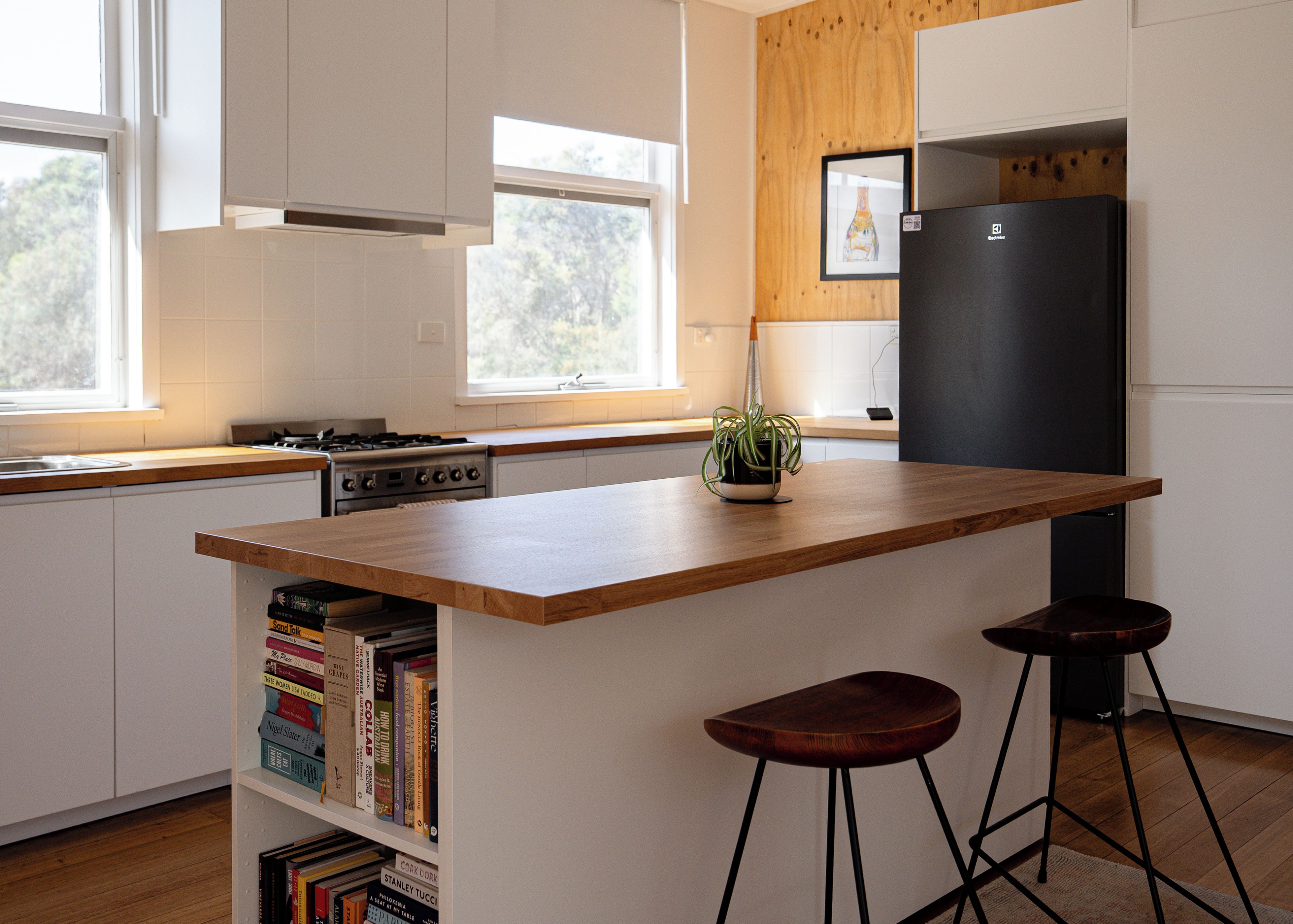 Une cuisine moderne avec des meubles blancs, un plan de travail en bois, un réfrigérateur noir et une bibliothèque remplie de livres.