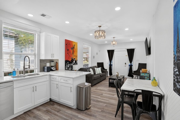 Una cocina moderna con armarios blancos, un comedor con mesa y sillas, y una sala de estar con sofá y televisor.