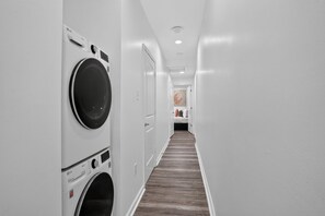 A hotel laundry room with two LG front-loading washing machines, a wooden floor, and a hallway leading to a bedroom with a bed and a painting. - Colorful New Orleans Home 3BR Near French Quarter (New Orleans)