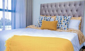 Una cama con cabecera capitonada, varios almohadones y una manta amarilla.