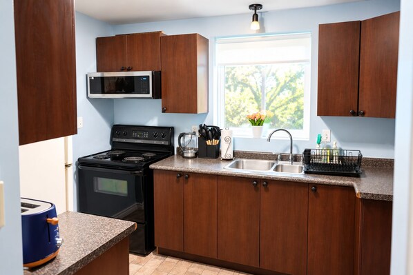 A kitchen with wooden cabinets, a stove, a microwave, a sink, and a window with a view of trees.